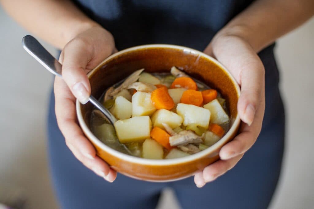 Tigela de sopa caseira de frango com cenoura e batata segurada com as mãos